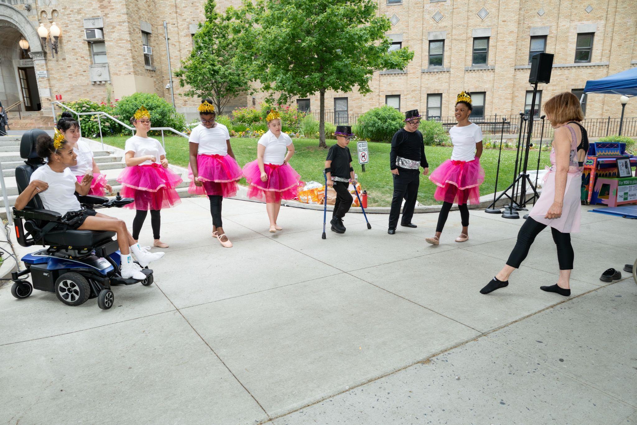 An adaptive dance performance with young performers in pink tutus and crowns, including a child in a power wheelchair and a child using crutches