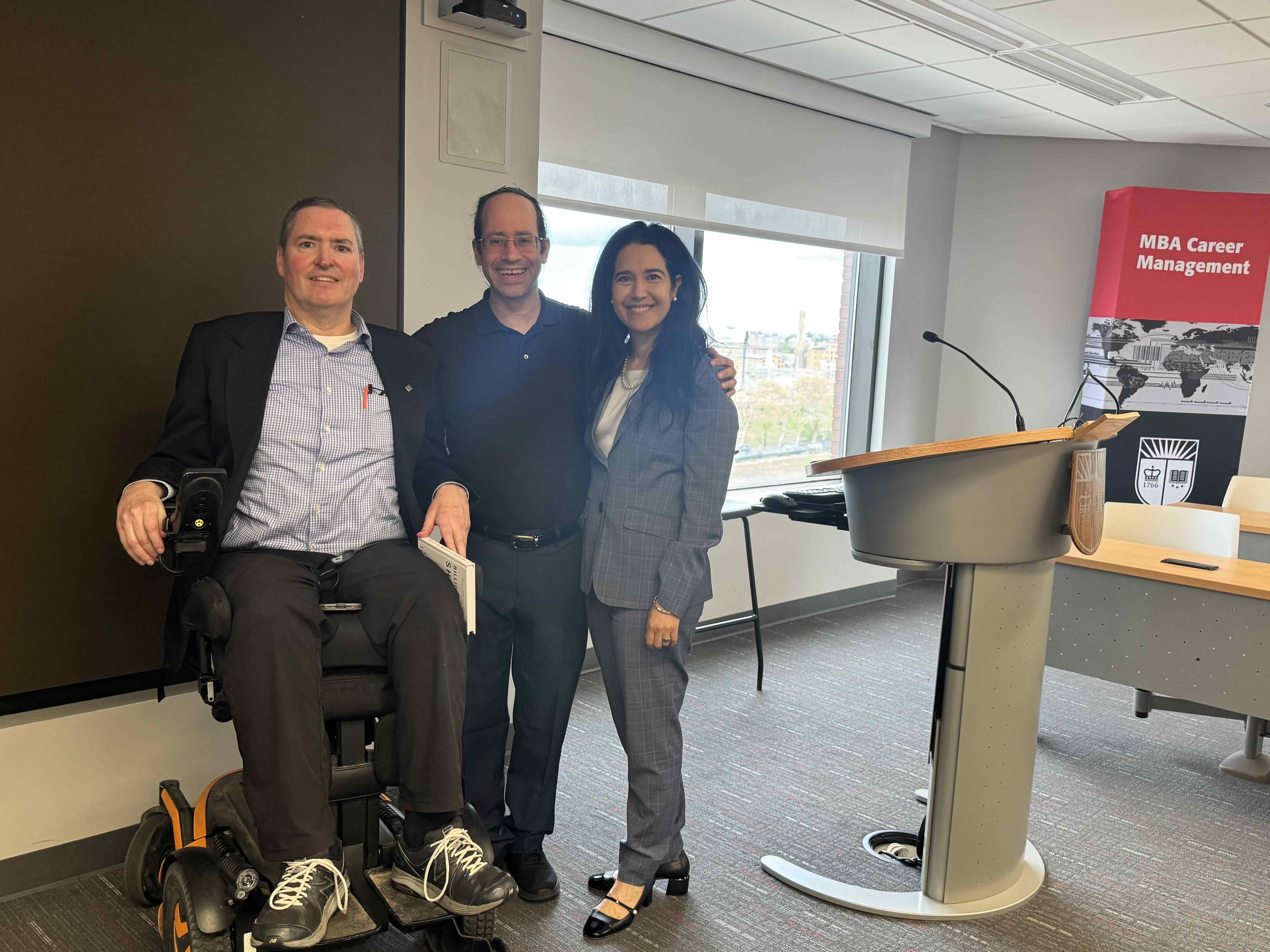 Three members of the Let's Move US team, one using a power wheelchair, standing together beside a lectern at the Rutgers Business School Business Plan Competition.